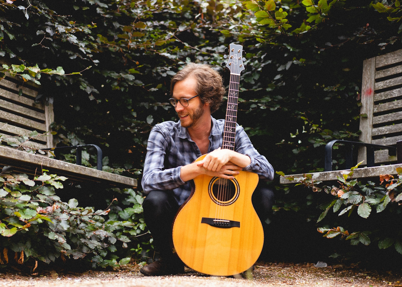 Danny Bradley crouching in a wooded area holding his guitar upright